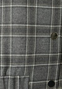 Tissu à carreaux gris avec des motifs noirs, blancs et un subtil orange. Doté d'un bouton en métal et d'une surface texturée.