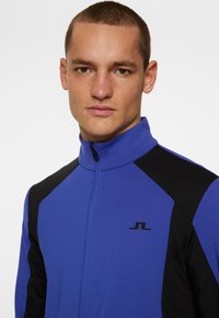 Blå och svart zip-jacka med hög krage, med en slät yta och en liten svart logotyp på bröstet.