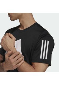 Camiseta atlética negra con un logo abstracto blanco en el pecho y tres rayas blancas en la manga izquierda, fabricada con un tejido suave.