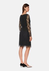 Schwarzes Spitzenkleid mit langen Ärmeln, tailliertem Schnitt und gewelltem Saum. Getragen mit zweifarbigen Schuhen, die ein Schwarz-Beige-Design aufweisen.