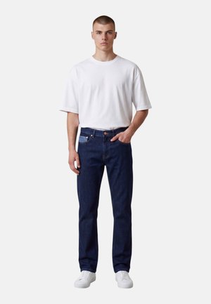 Giovane uomo in piedi rivolto in avanti che indossa una maglietta bianca semplice, jeans blu scuro e scarpe da ginnastica bianche su sfondo bianco.