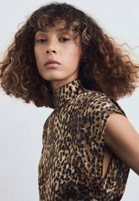 Mujer con cabello castaño rizado que lleva una blusa sin mangas de cuello alto con estampado de leopardo, mirando con confianza a la cámara frente a un fondo sencillo.