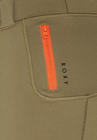 Los pantalones deportivos en verde oliva presentan un destacado bolsillo con cremallera naranja, con un logo cosido "ROXY" en negro y una textura suave.