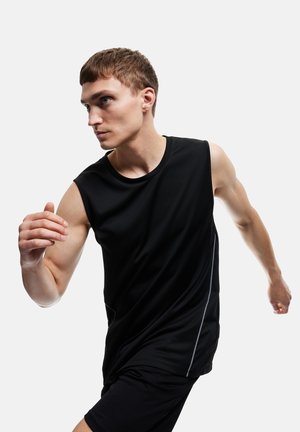 Joven con camiseta sin mangas negra y pantalones cortos corriendo, expresión concentrada, sobre fondo blanco liso.