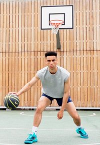 Baskettspiller som dribler en svart og gul ball. Han har på seg en grå ermeløs skjorte og blå shorts, med turkise treningssko.