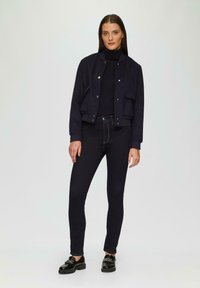 Marineblauwe jas met drukknoopsluitingen, voorzakken, gedragen over een marineblauwe coltrui; gecombineerd met donkere skinny jeans en zwarte loafers.