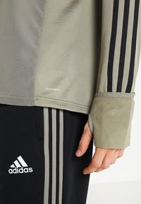 Beige langärmliges ClimaWarm-Oberteil mit schwarzen Streifen und Daumenloch, kombiniert mit schwarzen Adidas-Hosen mit grauen Streifen und Logo.