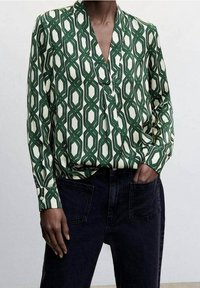 Groen en witte blouse met een V-hals, lange mouwen en een overlappend ontwerp aan de voorkant. Gecombineerd met donkere spijkerbroek.
