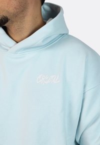 Felpa leggera di colore azzurro, realizzata in tessuto morbido, con una stampa grafica bianca della parola "CRYSTAL" sul petto. Include un cappuccio con corsa.
