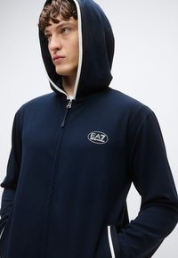 Sudadera con cremallera azul marino y capucha forrada de blanco, con un logo bordado en el pecho izquierdo y bolsillos laterales con detalles en blanco.