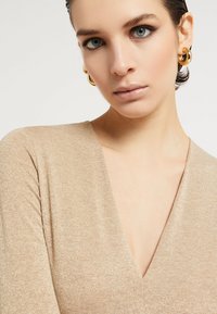 Bež top od pletenog materijala s sjajnim završetkom i dubokim V-izrezom. Glatka tekstura, prilagođen dizajn i vidljivi detalji šavova. Nošene su zlatne okrugle naušnice.