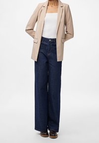 Blazer beige porté sur un top blanc ajusté, associé à un jean large bleu foncé et des chaussures marron. Le blazer présente des épaules structurées et des poches.