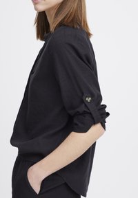 Camicia di lino nera con maniche lunghe, polsini arrotolati fissati da bottoni decorativi e vestibilità comoda. Tasche laterali visibili e tessuto morbido.