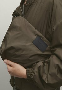 Olivgrüne Bomberjacke mit glatter Textur, Druckknopfverschluss und einem schwarzen Lederetikett auf der Vorderseite. Verfügt über elastische Bündchen.