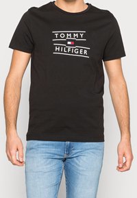 Hombre vistiendo una camiseta negra de Tommy Hilfiger con el logo y jeans de mezclilla azul claro, de pie contra un fondo plano.