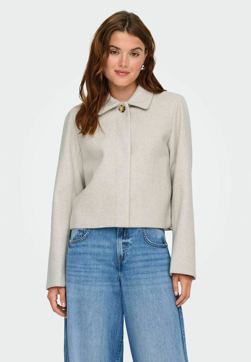 Femme portant une veste beige courte avec un col à un seul bouton et un jean bleu à jambes larges, sur un fond clair uni.