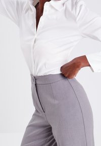 Chemise blanche boutonnée avec un col, associée à un pantalon gris clair ajusté avec une texture lisse et une taille cintrée.