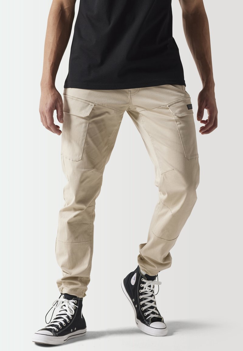 Pantalones cargo beige con múltiples bolsillos, textura suave, corte ajustado y costuras reforzadas. Combinados con zapatillas altas negras.