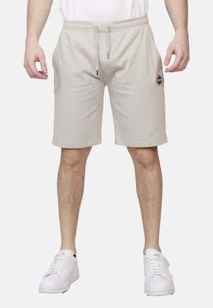 Uomo che indossa pantaloncini beige con coulisse, sneakers bianche e una camicia bianca, in piedi davanti a uno sfondo chiaro e uniforme.