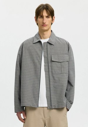 Młody mężczyzna w czarno-białej, w kratę kurtce na zamek, na białej koszuli i beżowych spodniach, stojący na jednolitym jasnym tle.