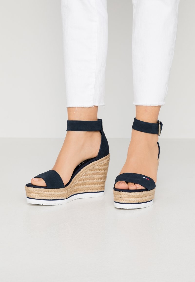 Pieds portant des sandales compensées bleu marine à bride cheville, talons enveloppés de jute et semelles blanches, assorties à un pantalon blanc court.