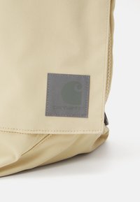 Béžový látkový batoh s texturovaným povrchem, šedou nášivkou loga a kapsou na zip. Design je jednoduchý a funkční.