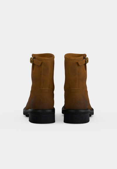 Paire de bottines en daim marron avec semelles noires et petites boucles latérales, vues de dos sur fond blanc.