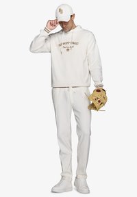Hvid hættetrøje med teksten "Varsity Team", matchende joggers, hvide sneakers og en baseballhandske i hånden, sat mod en ensfarvet baggrund.