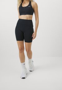 Brassière de sport noire avec fermeture éclair à l'avant et logo, associée à un short noir taille haute. Le mannequin porte des baskets blanches et des chaussettes avec un logo.