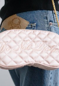 Vadderad rosa handväska med "Stör ej" broderat i ett slätt typsnitt. Väskan har ett guldkedjestropp och hålls nära denimjeans.