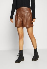 Pantalones cortos de cuero marrón con pliegues y una cinturilla alta, combinados con botas de tobillo negras con cordones. Textura suave y acabado elegante.