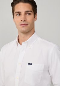 Camicia bianca con bottoni, dotata di una piccola tasca sul petto con etichetta blu, realizzata in un tessuto morbido, con colletto classico e dettagli di cucitura discreti.