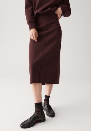 Mujer vistiendo una falda midi ajustada de color marrón oscuro, top de manga larga a juego, calcetines negros transparentes y mocasines de cuero negros contra un fondo liso.