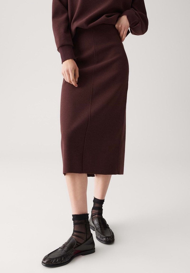 Femme portant une jupe midi marron foncé ajustée, un haut à manches longues assorti, des chaussettes noires transparentes et des mocassins en cuir noir devant un fond uni.