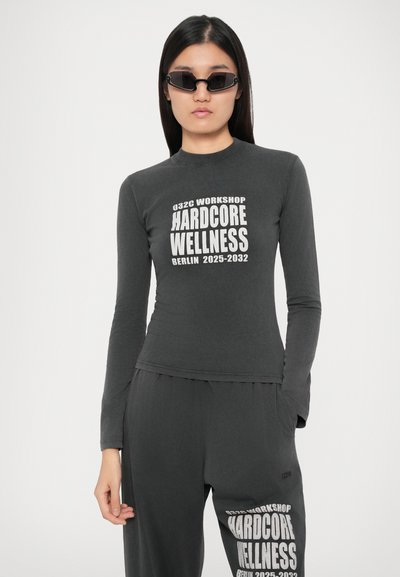 Žena v tmavosivej dlhý rukáv top a nohaviciach s textom "032C Workshop Hardcore Wellness Berlin 2025-2032" a čiernych úzkych slnečných okuliaroch.