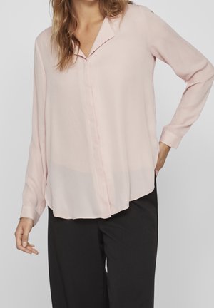 Femme portant un chemisier rose clair à manches longues et un pantalon noir, posant avec une main sur la hanche et l'autre détendue le long du corps.