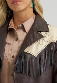 Smeđa kožna jakna s kremastim naglascima, resama i ovratnikom s prorezom. Nosi se preko svijetloružičaste košulje s gumbima.