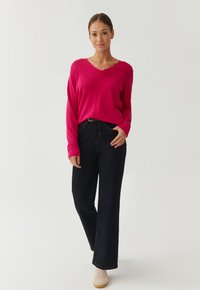 Fuchsia V-Ausschnitt Pullover kombiniert mit dunkelblauen weit geschnittenen Jeans und hellen Sneakers. Glatte Textur, lässige Passform.