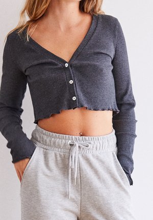 Donna che indossa un top corto grigio scuro a coste con maniche lunghe e pantaloni tuta grigio chiaro con cordino, con le mani nelle tasche.