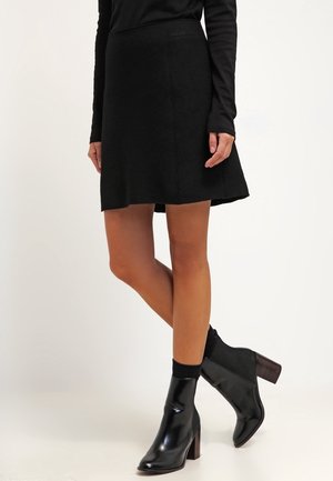 Personne portant un haut noir à manches longues, une jupe trapèze noire et des bottines noires brillantes à talons blocs sur un fond uni.