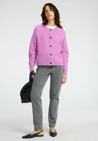 Rosa, gestrickte Strickjacke mit Rundhalsausschnitt und schwarzen Knöpfen, kombiniert mit grauen Jeans und schwarzen Slingback-Ballerinas. Einheitlicher Hintergrund.