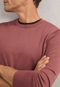 Koszulka z długim rękawem i okrągłym dekoltem w stonowanym kolorze róży, z teksturowanym wzorem dzianiny. Podszewka widoczna w ciemnym bordowym kolorze.