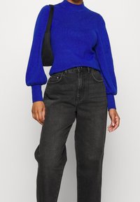 Maglione blu lavorato a maglia con collo alto e maniche a sbuffo abbinato a jeans neri a vita alta. Borsa nera indossata sulla spalla.