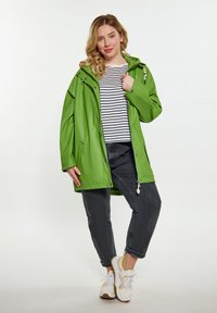 Žalias vandeniui atsparus švarkas su užtrauktuku, dideliu gobtuvu ir dviem šoniniais kišenėmis, vilkimas virš juostele išmarginto marškinėlių ir juodų kelnių, su baltais sportbačiais.