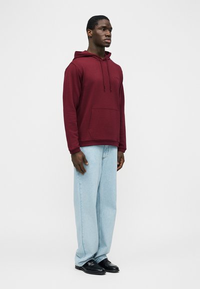 Άνδρας στέκεται φορώντας μπορντό φούτερ με κουκούλα, ανοιχτό μπλε τζιν με φαρδιά παντελόνια και μαύρα παπούτσια μπροστά σε απλό λευκό φόντο.