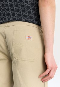 Beige Dickies bukser med en baglomme med et logo patch, glat tekstur og lige ben design, vist fra en sidevinkel.