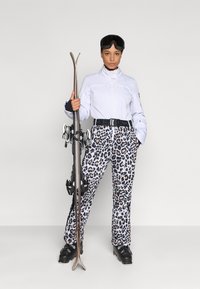 Costum de schi compus dintr-o jachetă albă ușoară și pantaloni cu imprimeu de leopard. Accesoriile includ o curea de schi neagră și ochelari de schi, ținând schiuri.