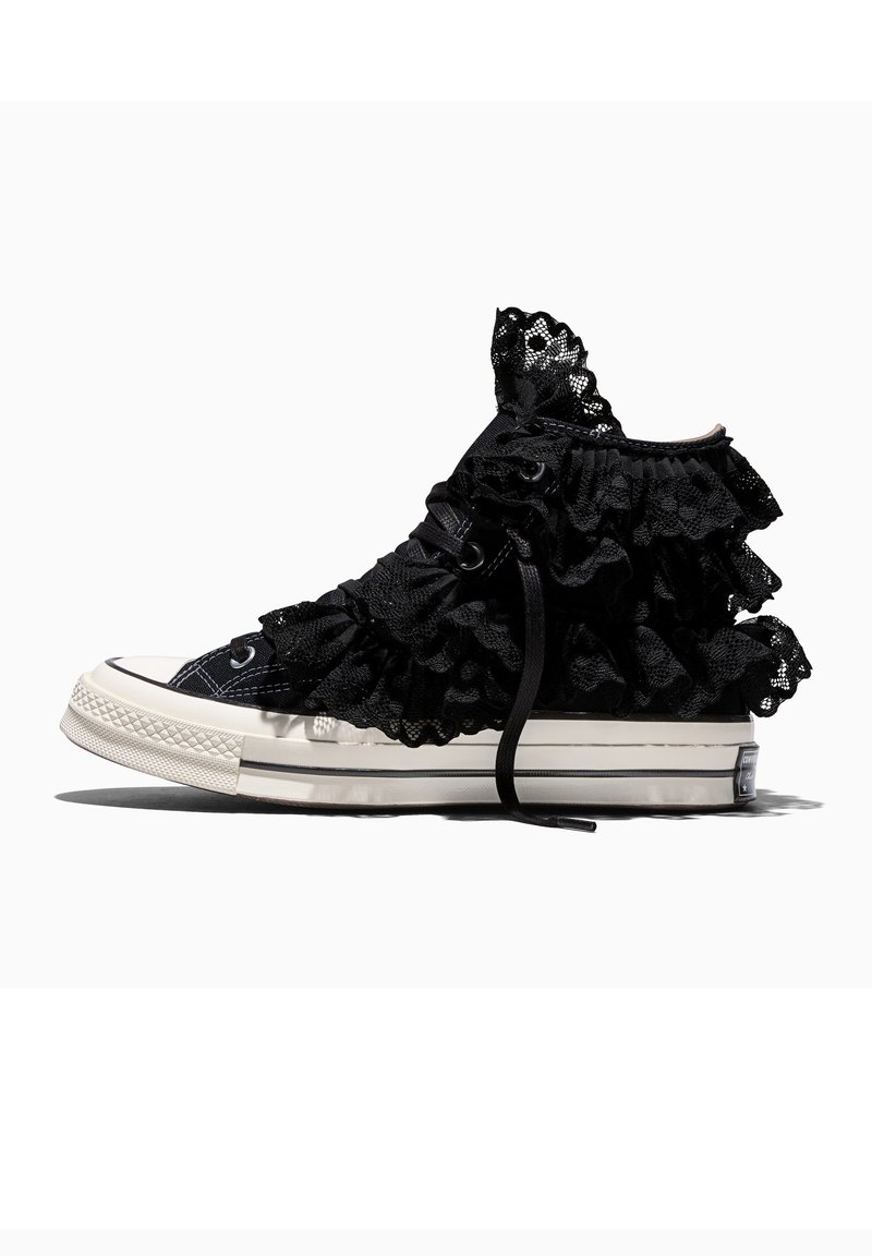 Zapatilla negra de caña alta con suela blanca, adornada con múltiples capas de volantes de encaje negro alrededor del tobillo y los lados superiores.