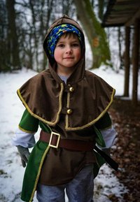 Brown and green hooded cloak with gold trim and buttons, worn over a gray shirt. Blue-patterned hat and gray gloves complete the outfit. Snowy background.