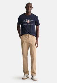 T-shirt bleu marine avec logo blanc et rouge ; pantalon en coton beige ; baskets blanches avec accents noirs. Look décontracté et sportif simple.